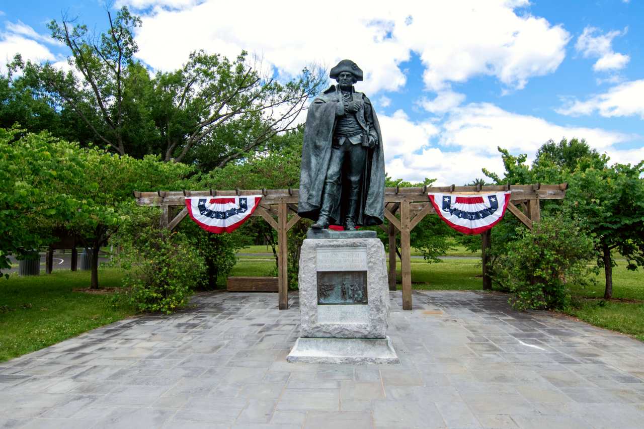 bronze statue of rev war general on stone base, red white and blue bunting behind