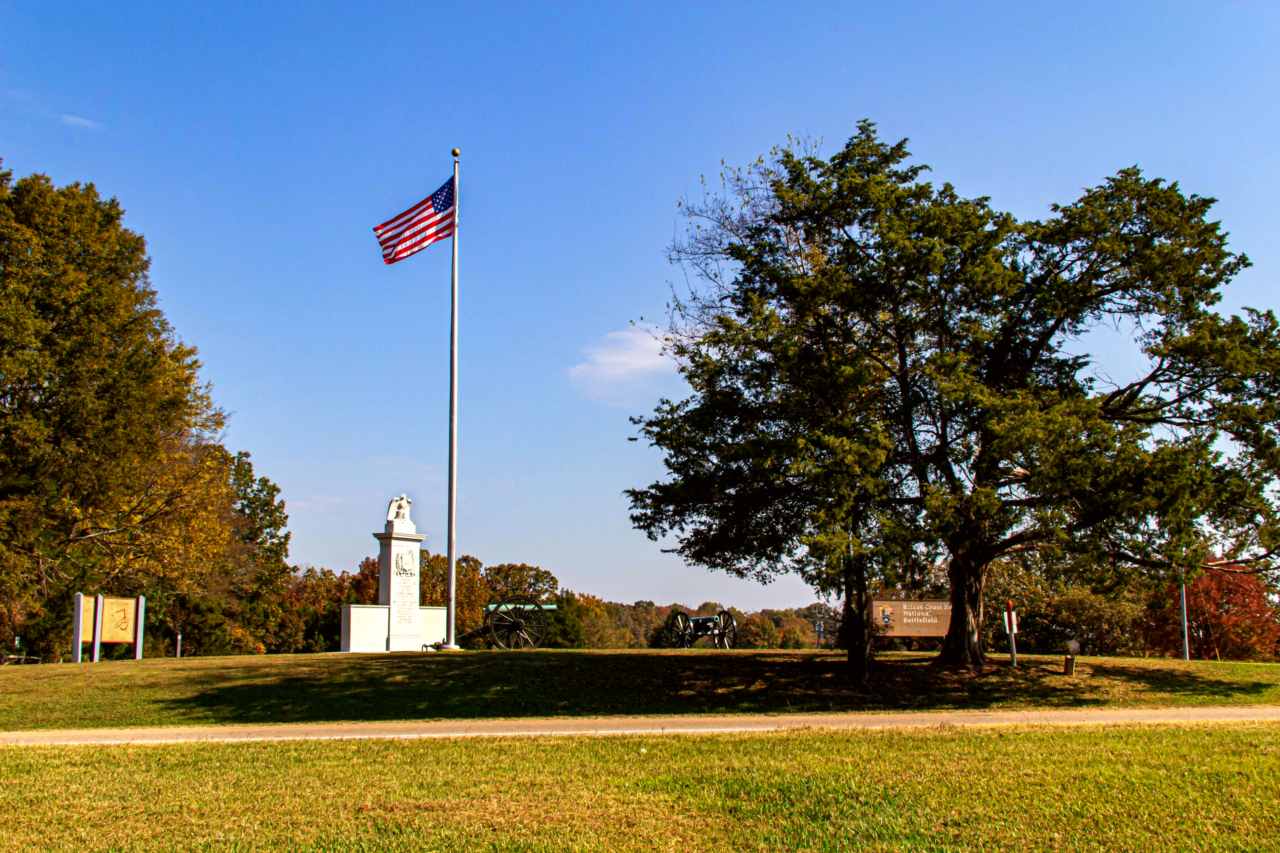 Two large trees and a flagpole flying an American flag overlooking a stone monument and cannons