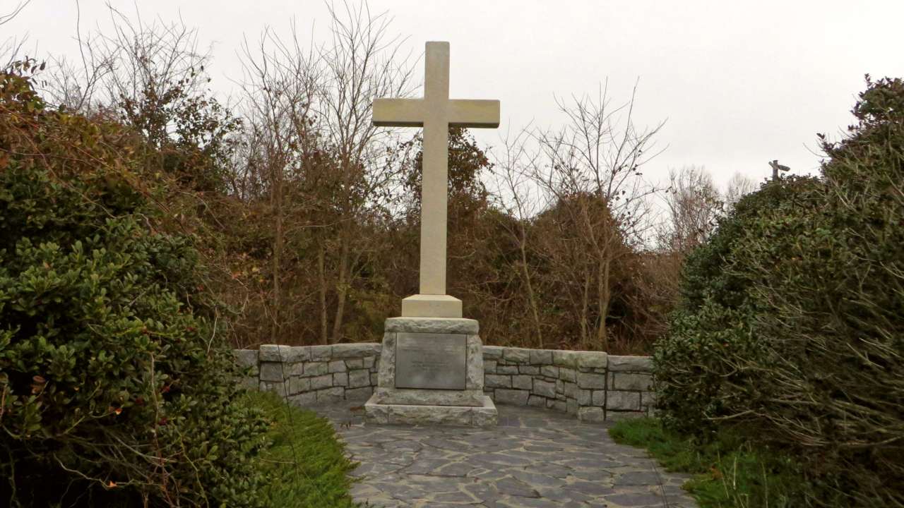 Gray stone cross monument in front of a low stone wall at the end of a flagstone path