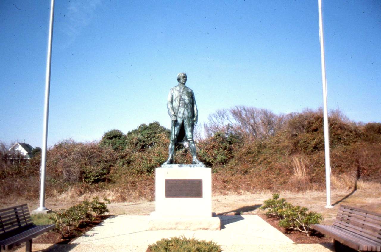 Patinaed statue of a military officer flanked by flagpoles and benches
