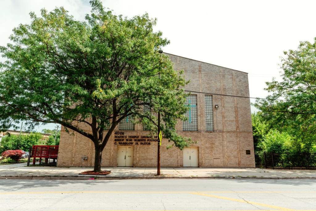 A multi-story tan brick church along a road. A tree stands in front.