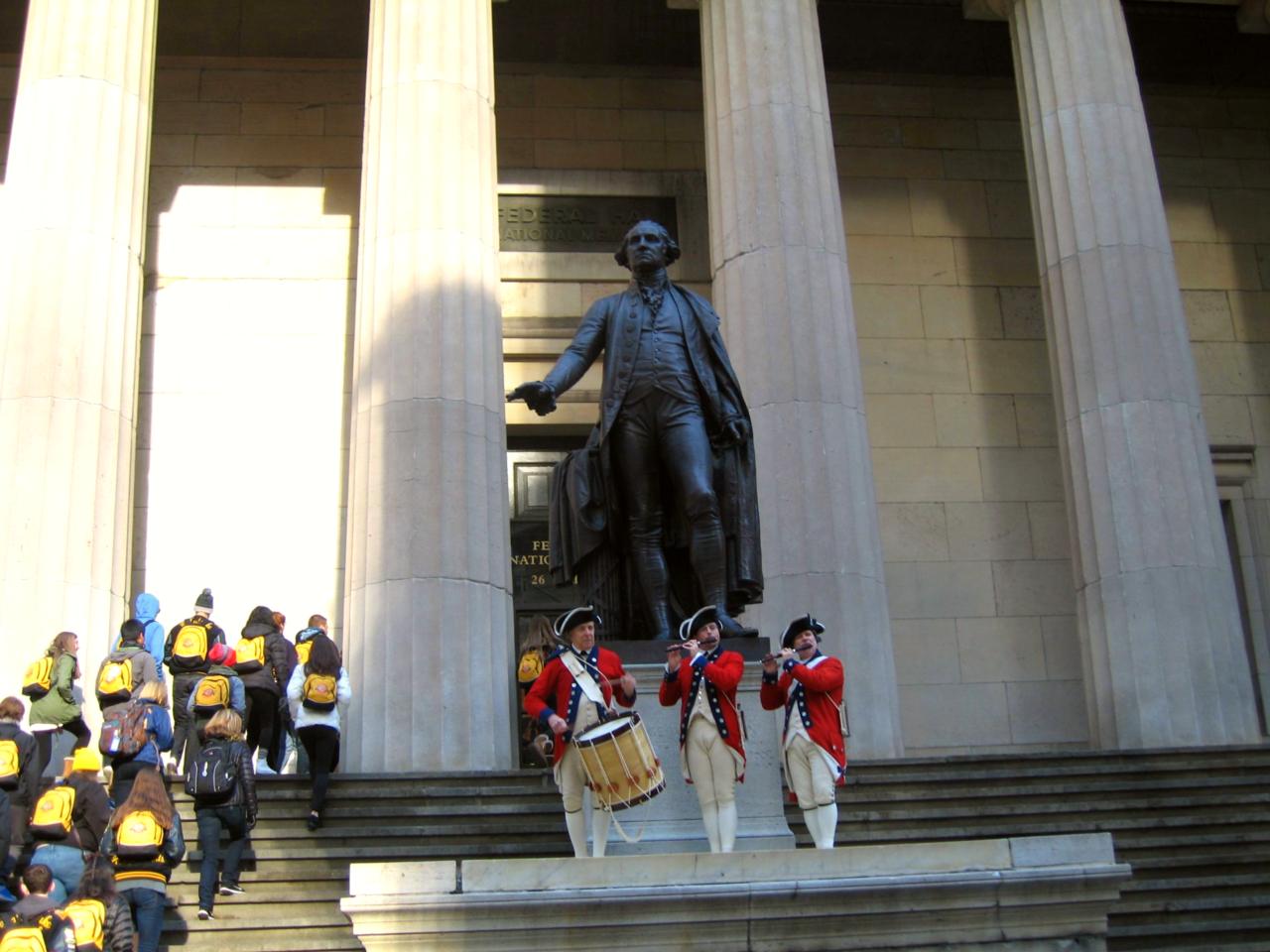 Three people in reenactment clothing hold instruments in front of a stone building and giant statue.