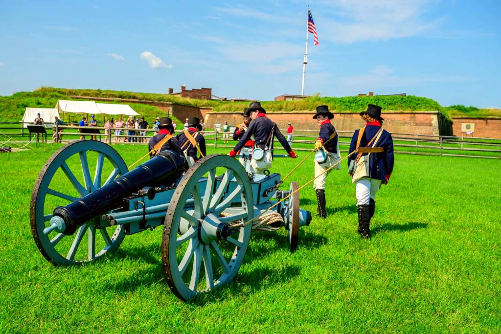 Men in War of 1812 era uniforms pull a cannon over grass outside of a brick fort.