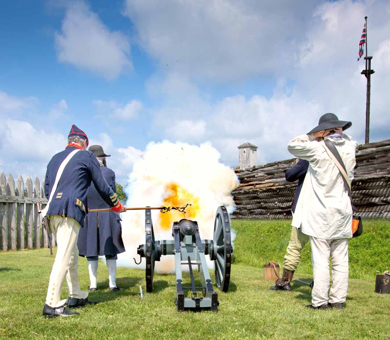 A cannon crew fires towards a wooden fort. An explosion of fire jets out in front of them.