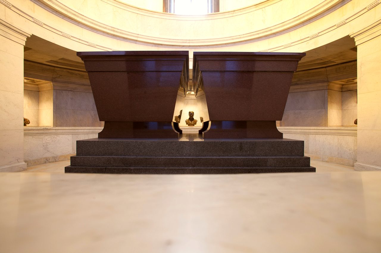 Two red stone sarcophagi are in a crypt. A bust of General Sherman can be seen in the distance.