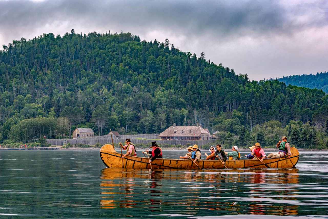 People in historic clothing paddle a canoe in a bay in front of a stockade.