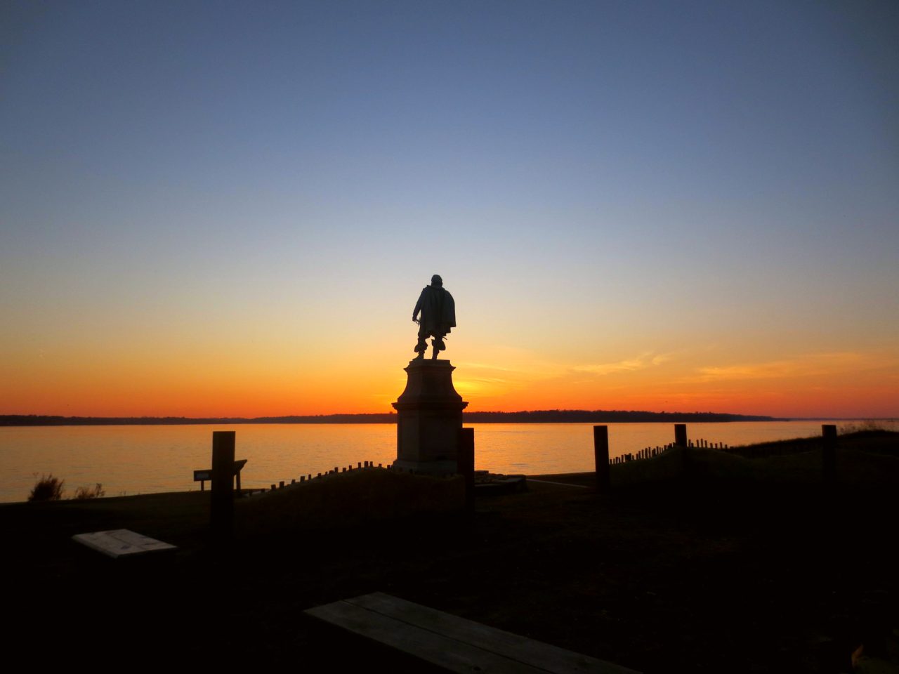 Statue on a pedestal silhouetted against a sunset over a river