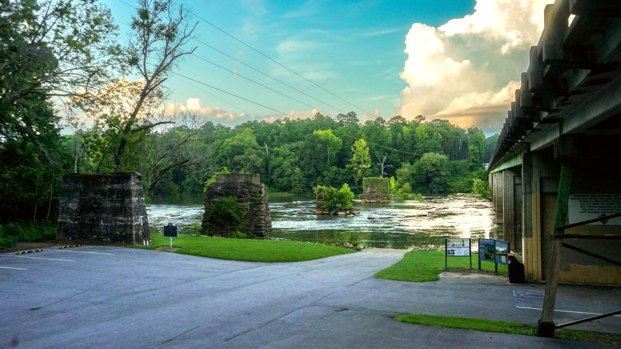 flat river, green trees lining banks, old bridge pier made of stones in river on left side of photo