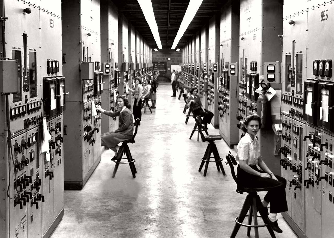 Several women sit on stools along a long hallway full of dials and meters.