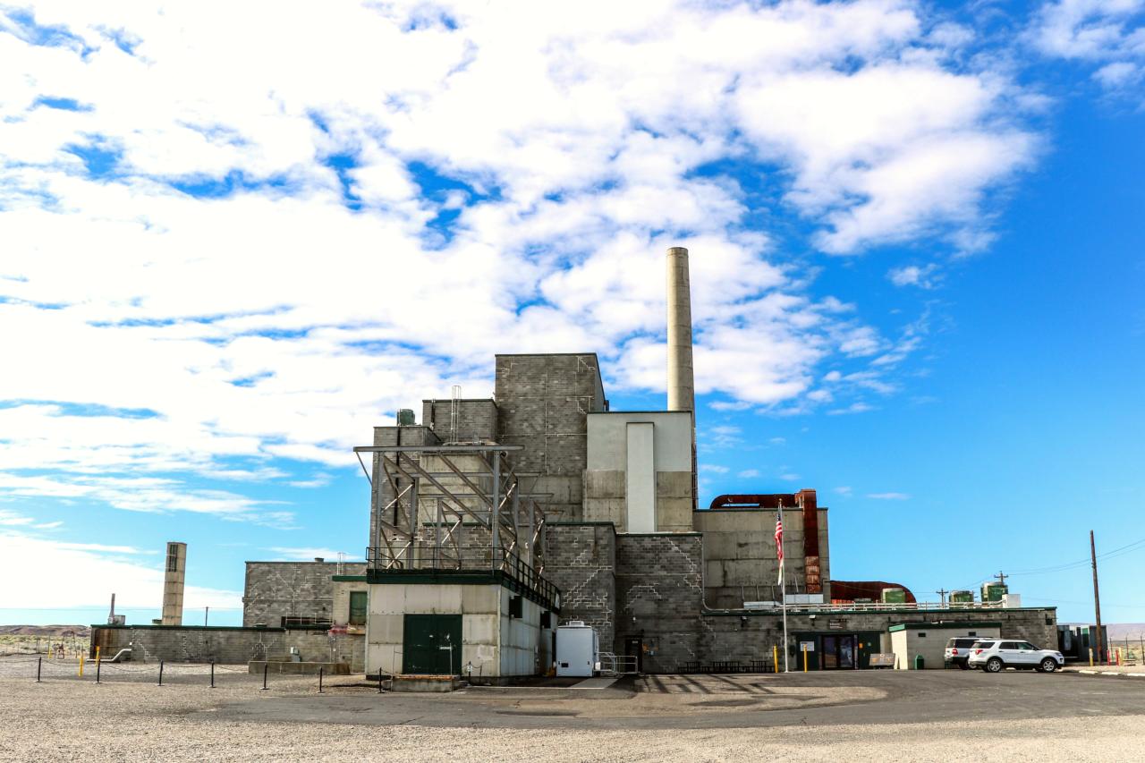 Gray factory building like stacked boxes with single taller smokestack beneath cloudy blue skies.