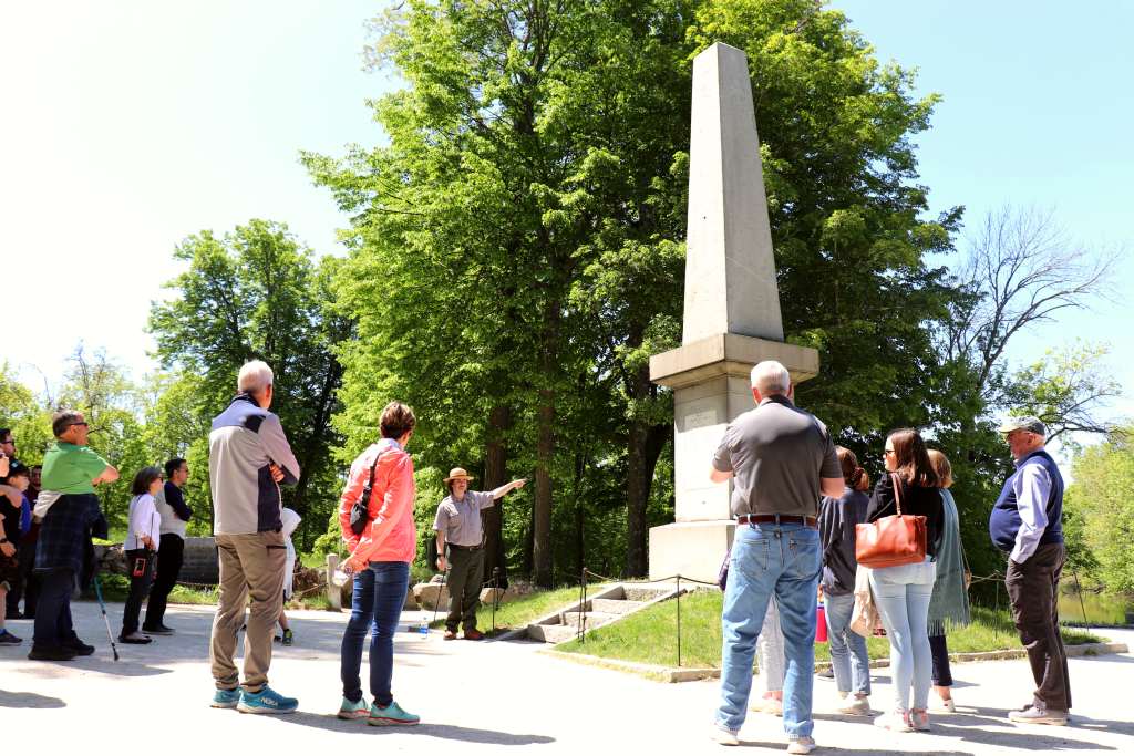 A park ranger stands in front of a stone obelisk monument pointing and talking to visitors