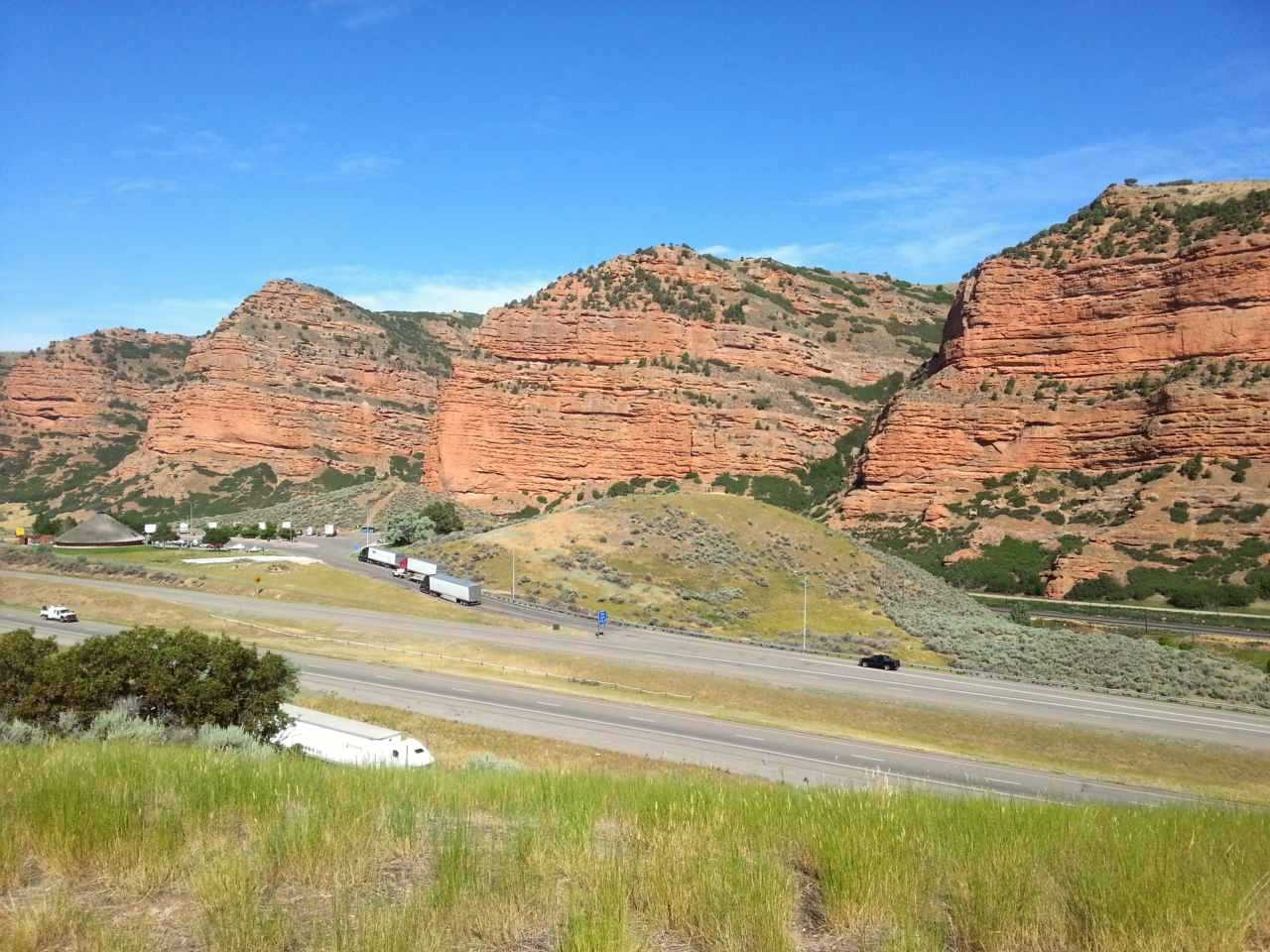 Red cliffs line a highway as seen from a high point with grass in the foreground.