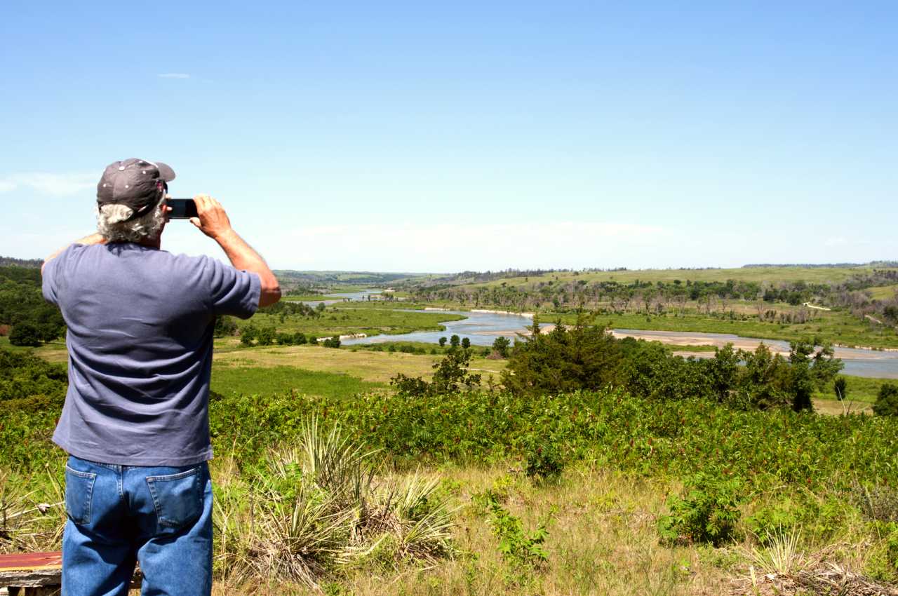 A man takes a photo of a meandering river from an overlook.