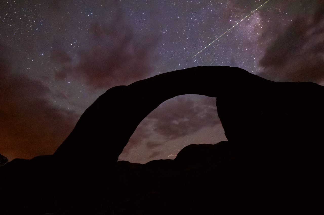 A silhouetted rock arch under a stary sky with a satellite flying by.