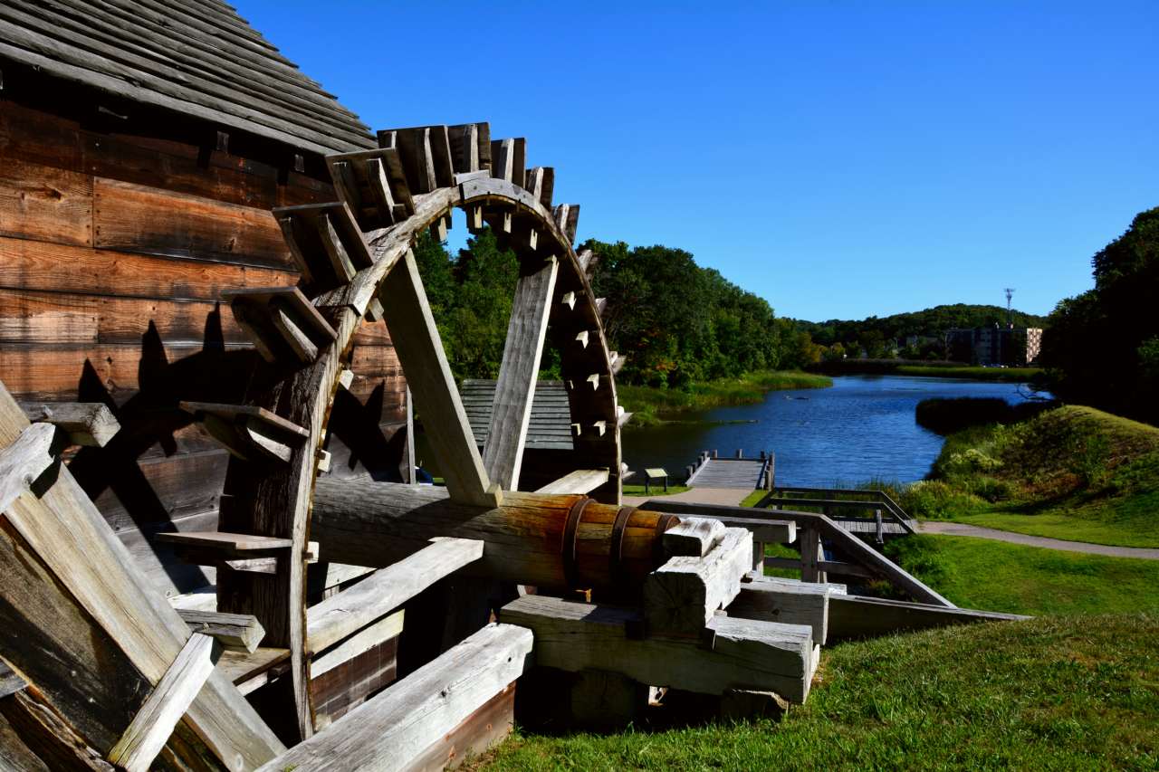 Approximately 15' by 15' spoked wooden wheel beside river and under blue sky.