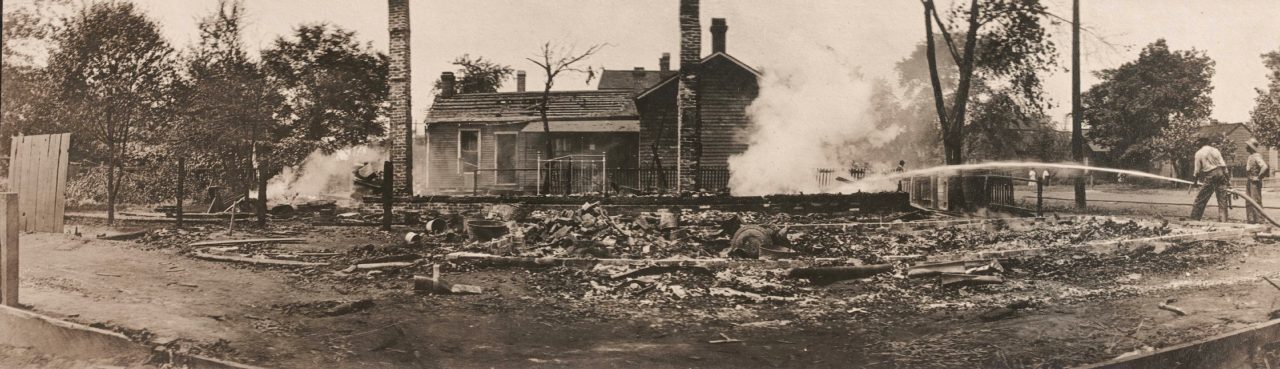 Historic photo of two people using a hose to extinguish a burning building.