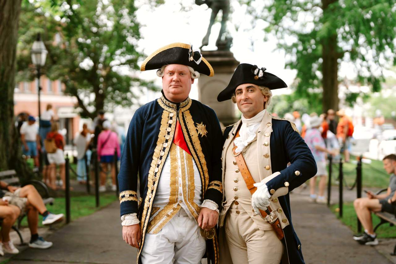Two reenactors in Revolutionary War-era military uniforms stand together in a park.