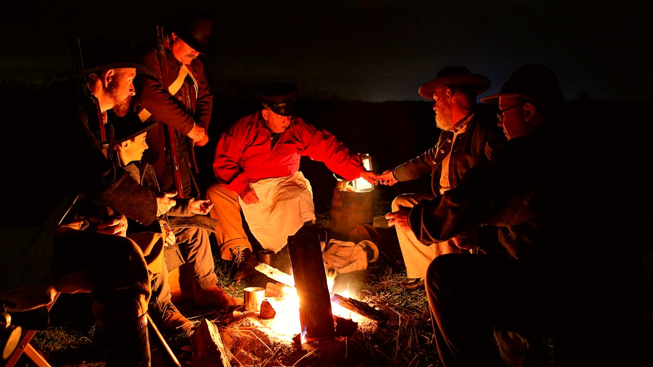 Men in reproduction uniforms talk around a campfire