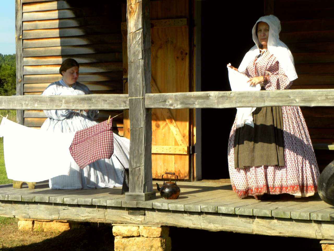 Women in reproduction historic dress sit and stand on porch of old cabin