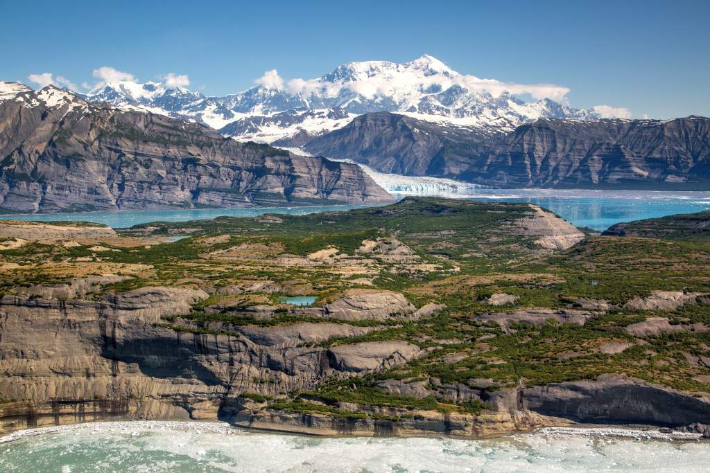 Glaciers loom over the ocean with large snowy mountains rising into blue skies