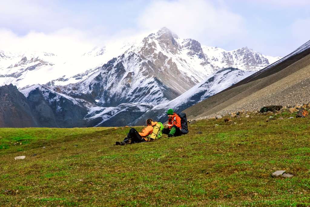 Two backpackers sitting in an alpine meadow with snowy mountains in the background