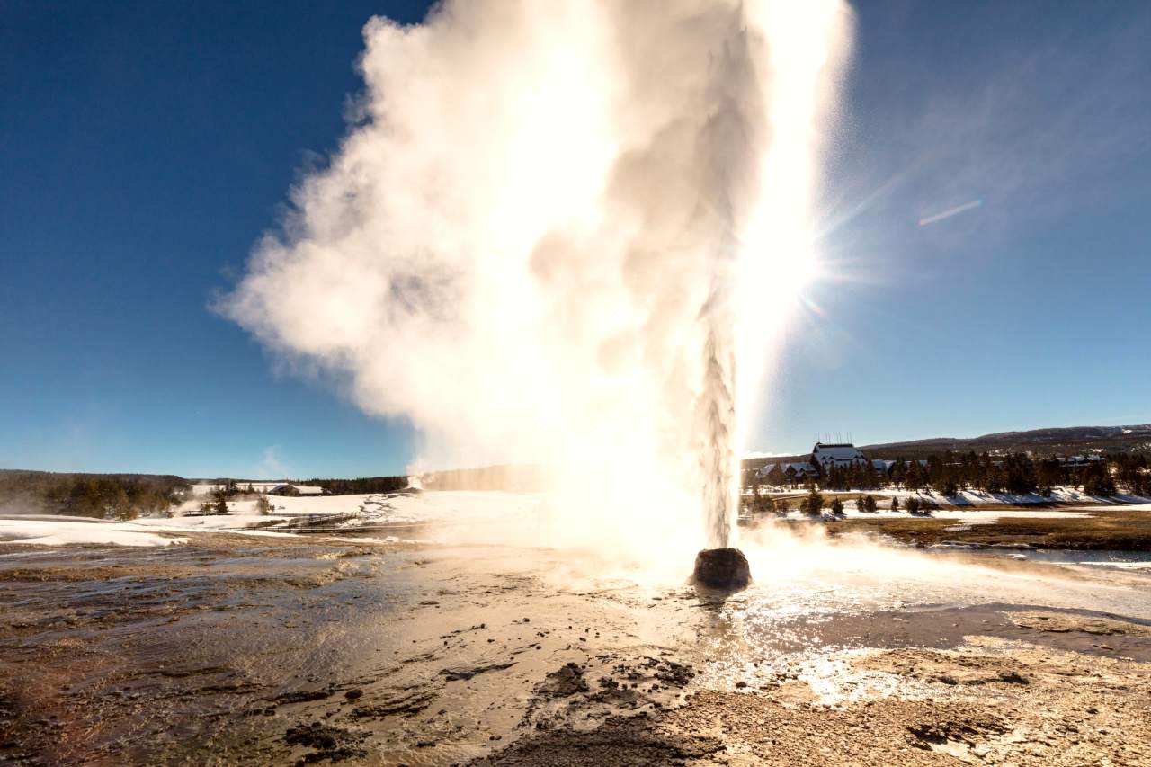 A geyser erupts steam in front of the bright sun.