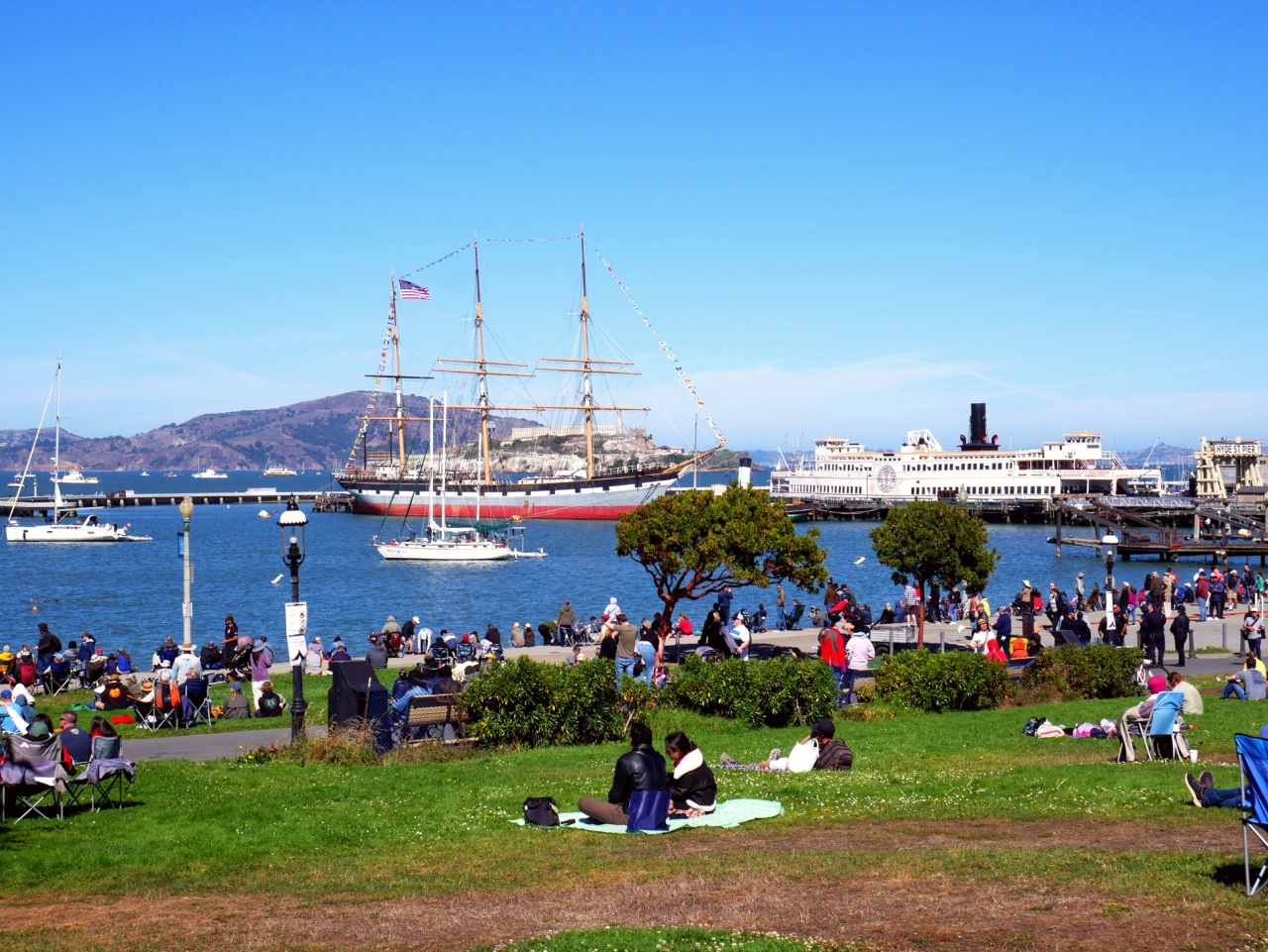 People sitting on a grassy lawn look out over a cove and historic ships in the distance