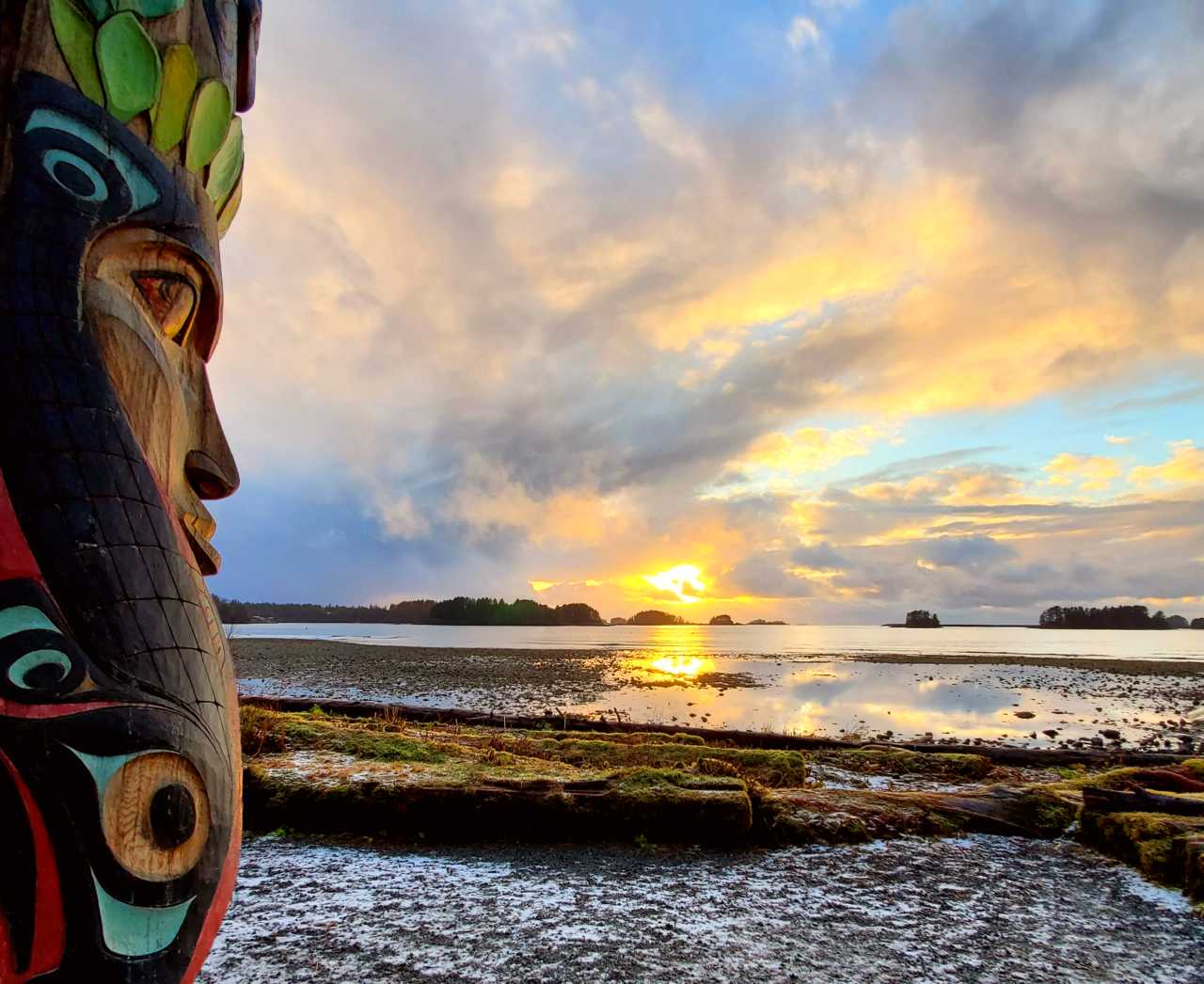 A totem pole in the foreground with a sunset over water in the background.
