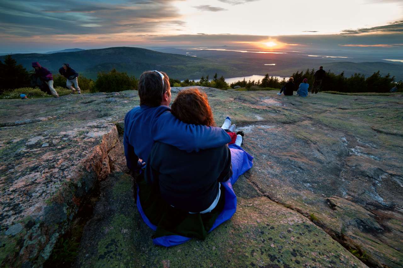 While seated on a blanket on a patch of mountaintop granite, a man and woman clasp their arms around