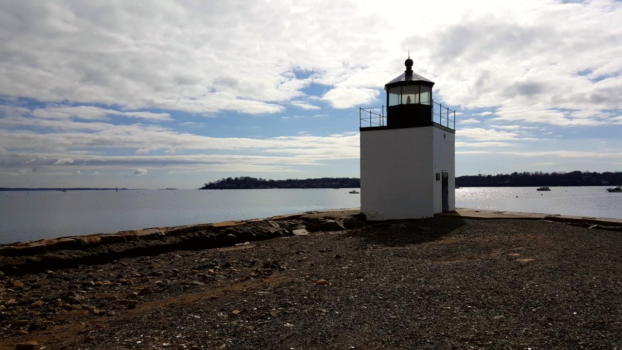 A white rectangular lighthouse approximately 20 feet tall is near the water on a gravel road.