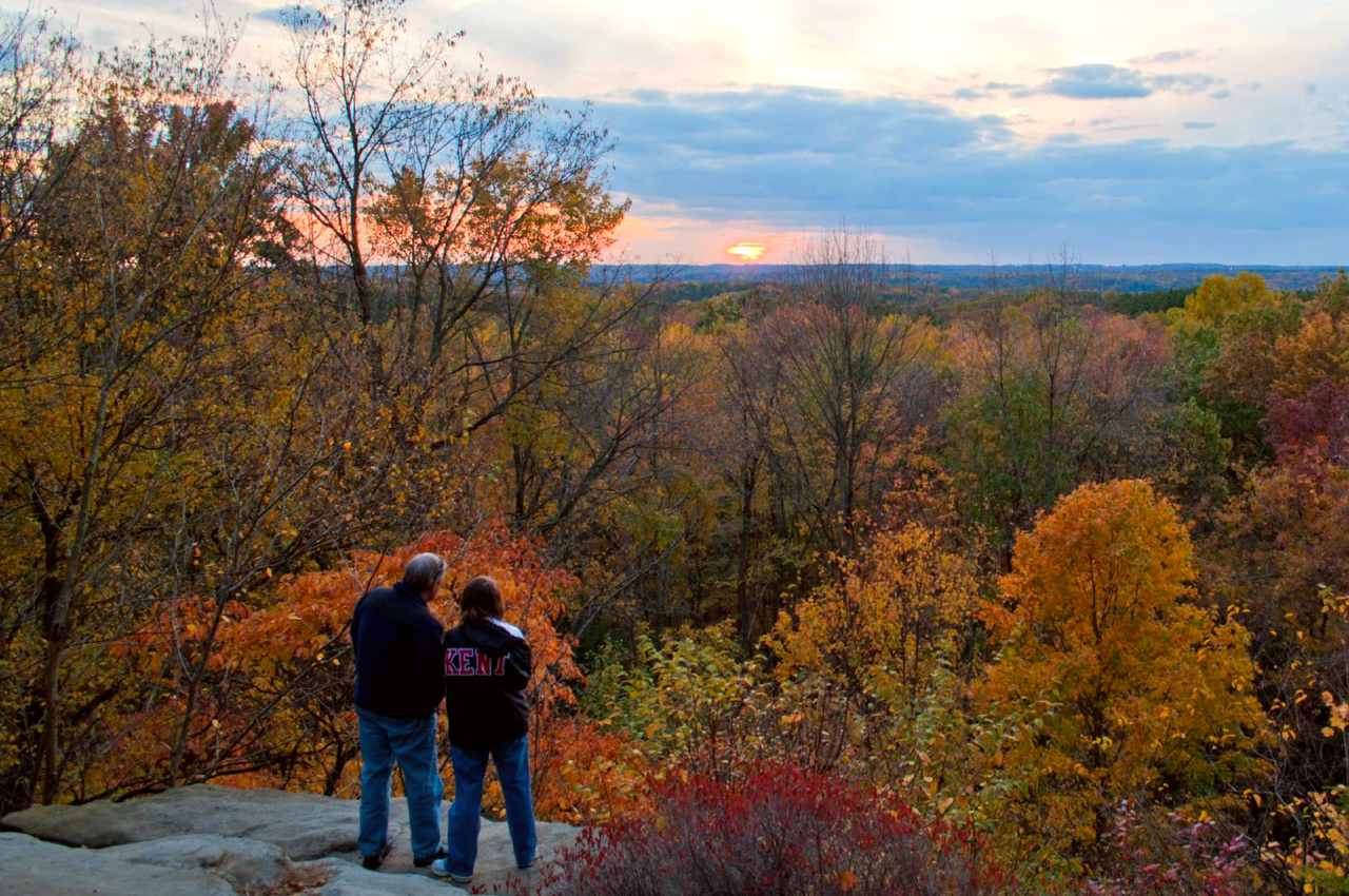 Two people stand at the edge of a gray rock cliff; in the distance, sunset and trees in fall colors.